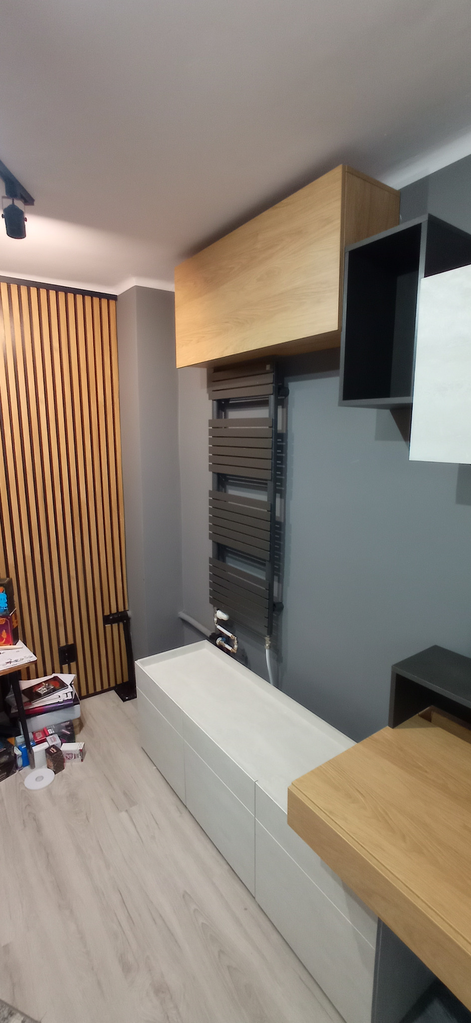 Szary grzejnik drabinkowy zamontowany na ścianie nad białą szafką RTV, obok półki z czarnym elementem i drewnianej szafki wiszącej, na podłodze panele, na ścianie panele dekoracyjne.