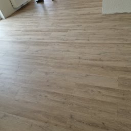 Parkett Jakubowski - Jasne panele podłogowe z widocznym fragmentem pracownika w stroju roboczym, częściowo ułożone w pustym pomieszczeniu z białymi ścianami i grzejnikiem.