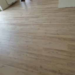 Jasne panele podłogowe z widocznym fragmentem pracownika w stroju roboczym, częściowo ułożone w pustym pomieszczeniu z białymi ścianami i grzejnikiem.