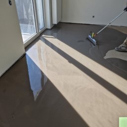 Parkett Jakubowski - Równomierne rozprowadzanie wylewki samopoziomującej na podłodze w jasnym pomieszczeniu z dużym oknem balkonowym, widoczne narzędzie do rozprowadzania i fragment buta pracownika.