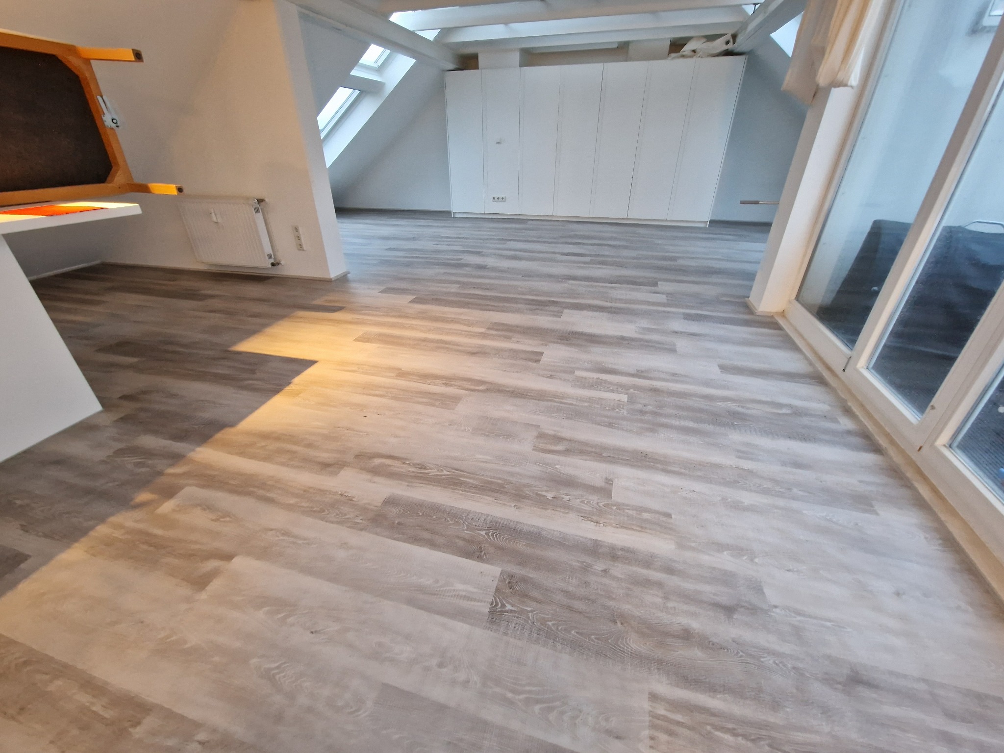 Jasna, przestronna mansarda z nową podłogą z paneli w odcieniach szarości, imitujących drewno, widoczne skosy, okno dachowe i biała zabudowa meblowa.