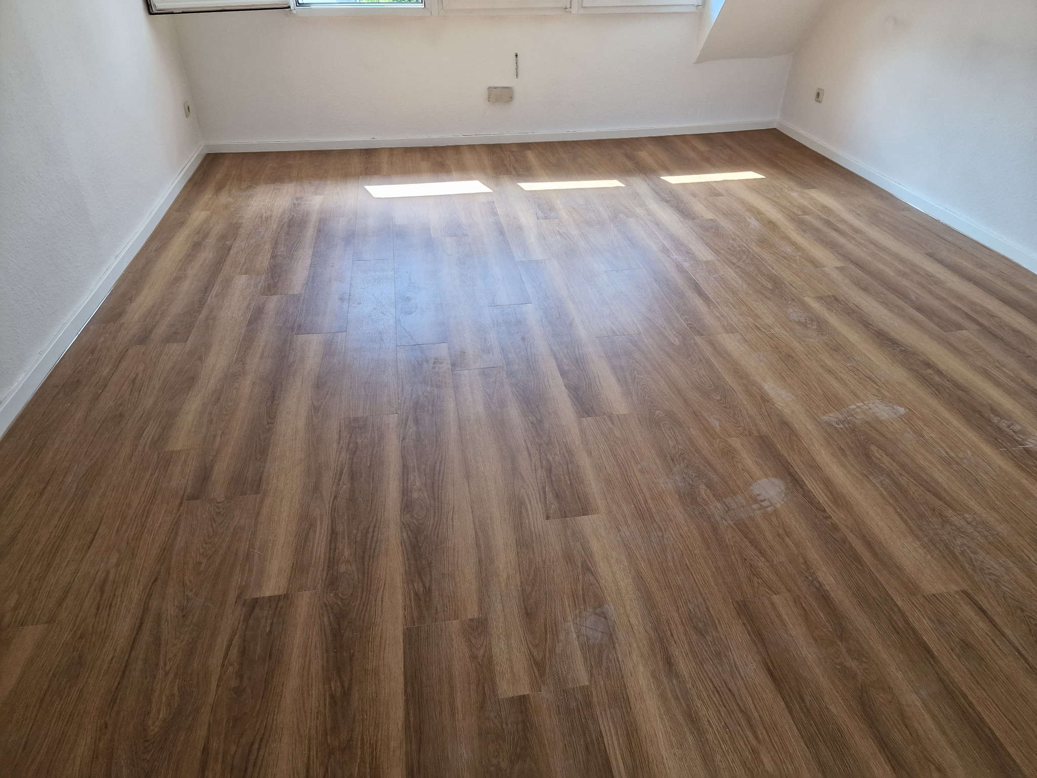 Nowo ułożona, brązowa podłoga z paneli laminowanych w pustym pokoju na poddaszu, z widocznymi promieniami słonecznymi wpadającymi przez okno dachowe.