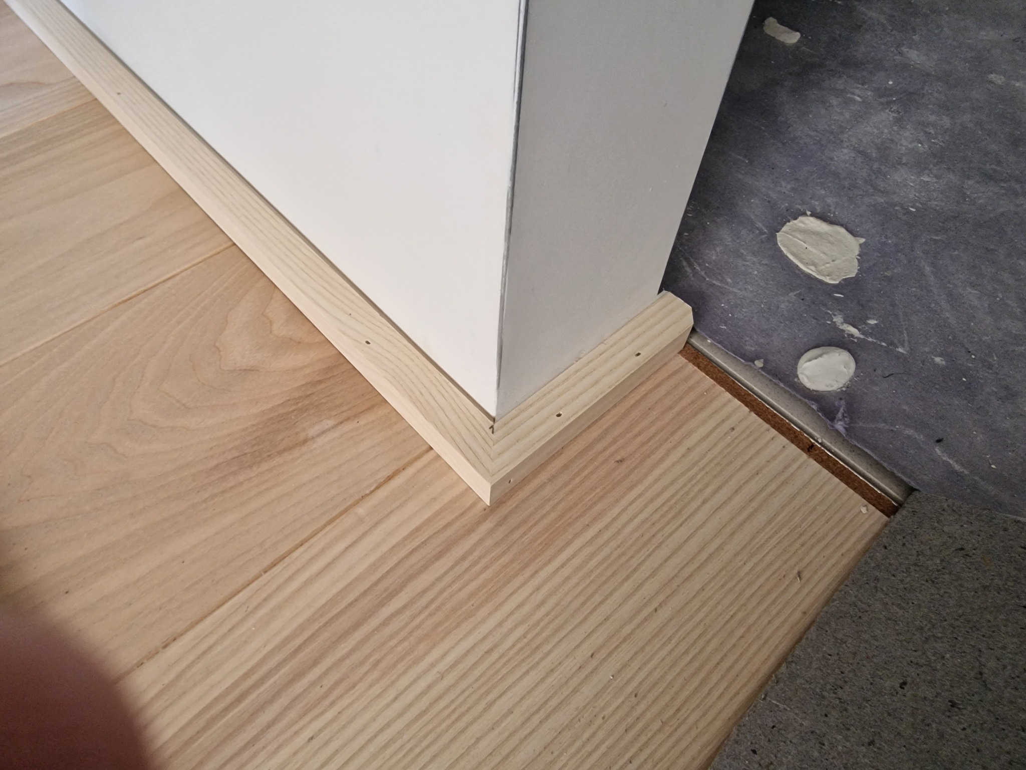 Fragment podłogi z jasnych paneli drewnianych, widoczne listwy przypodłogowe z surowego drewna przy białej ścianie i fragment szarej podłogi z plamami zaprawy.