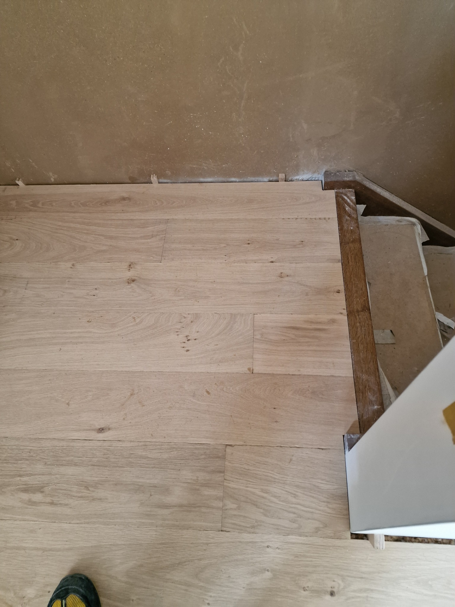 Jasne panele podłogowe ułożone wzdłuż ściany z widocznymi szczelinami i listwami przypodłogowymi, fragment schodów w tle.