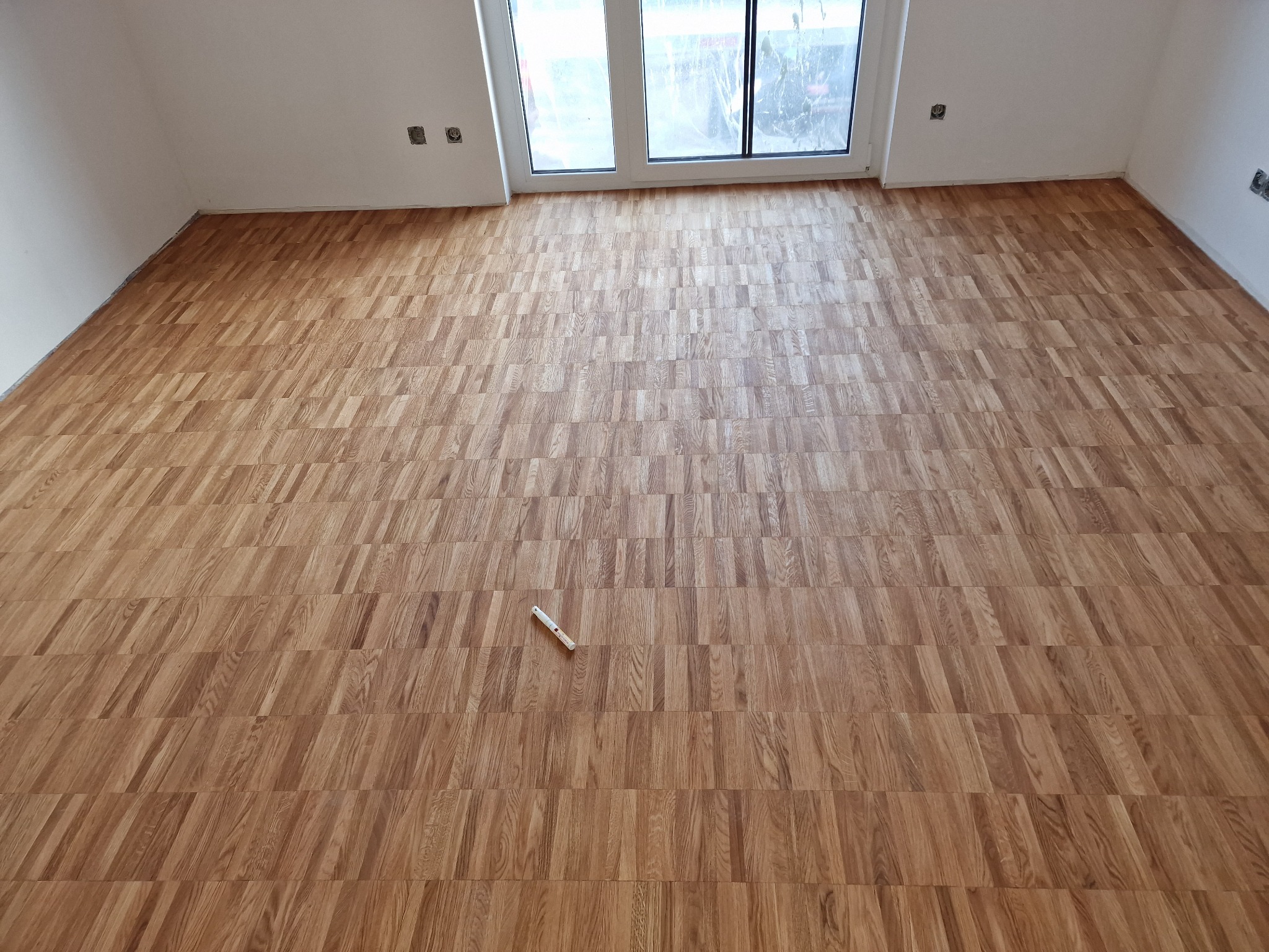 Ułożony parkiet w pustym pokoju z białymi ścianami i oknem balkonowym, widoczny biały przyrząd do pomiaru na podłodze.