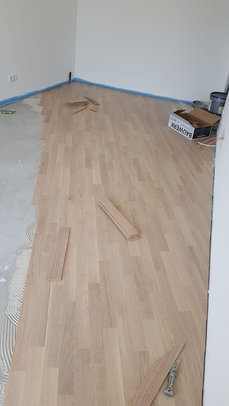 Układanie jasnego parkietu w pomieszczeniu, widoczne fragmenty nieułożone, paczka paneli Bauwerk Mondpark, narzędzia i klej.