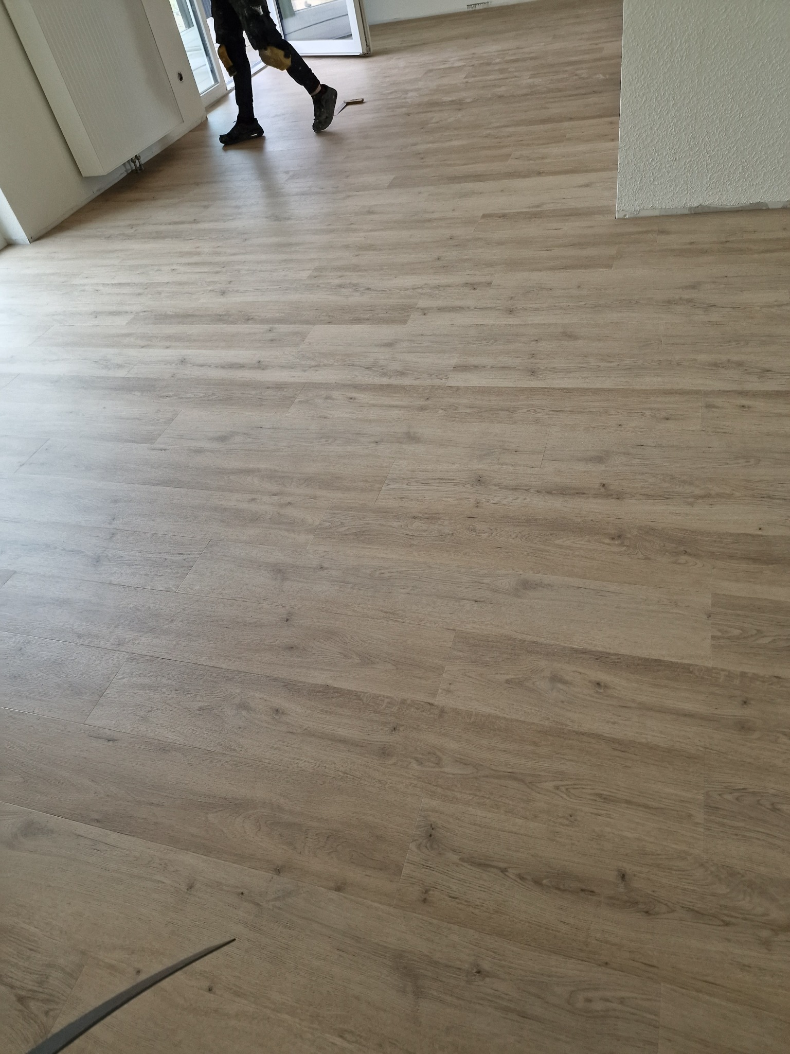 Jasne panele podłogowe z widocznym fragmentem pracownika w stroju roboczym, częściowo ułożone w pustym pomieszczeniu z białymi ścianami i grzejnikiem.