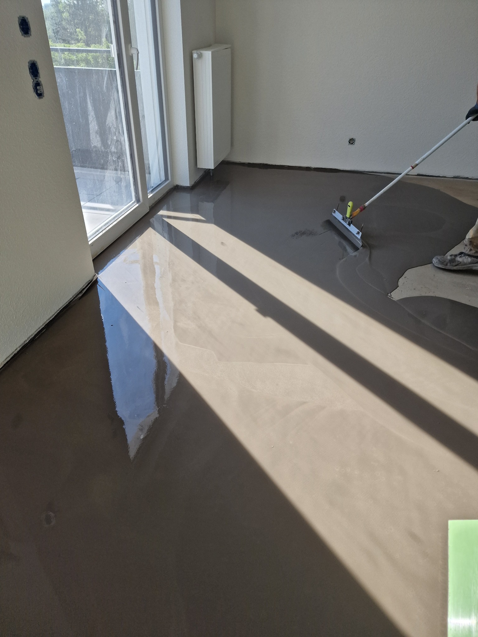Równomierne rozprowadzanie wylewki samopoziomującej na podłodze w jasnym pomieszczeniu z dużym oknem balkonowym, widoczne narzędzie do rozprowadzania i fragment buta pracownika.