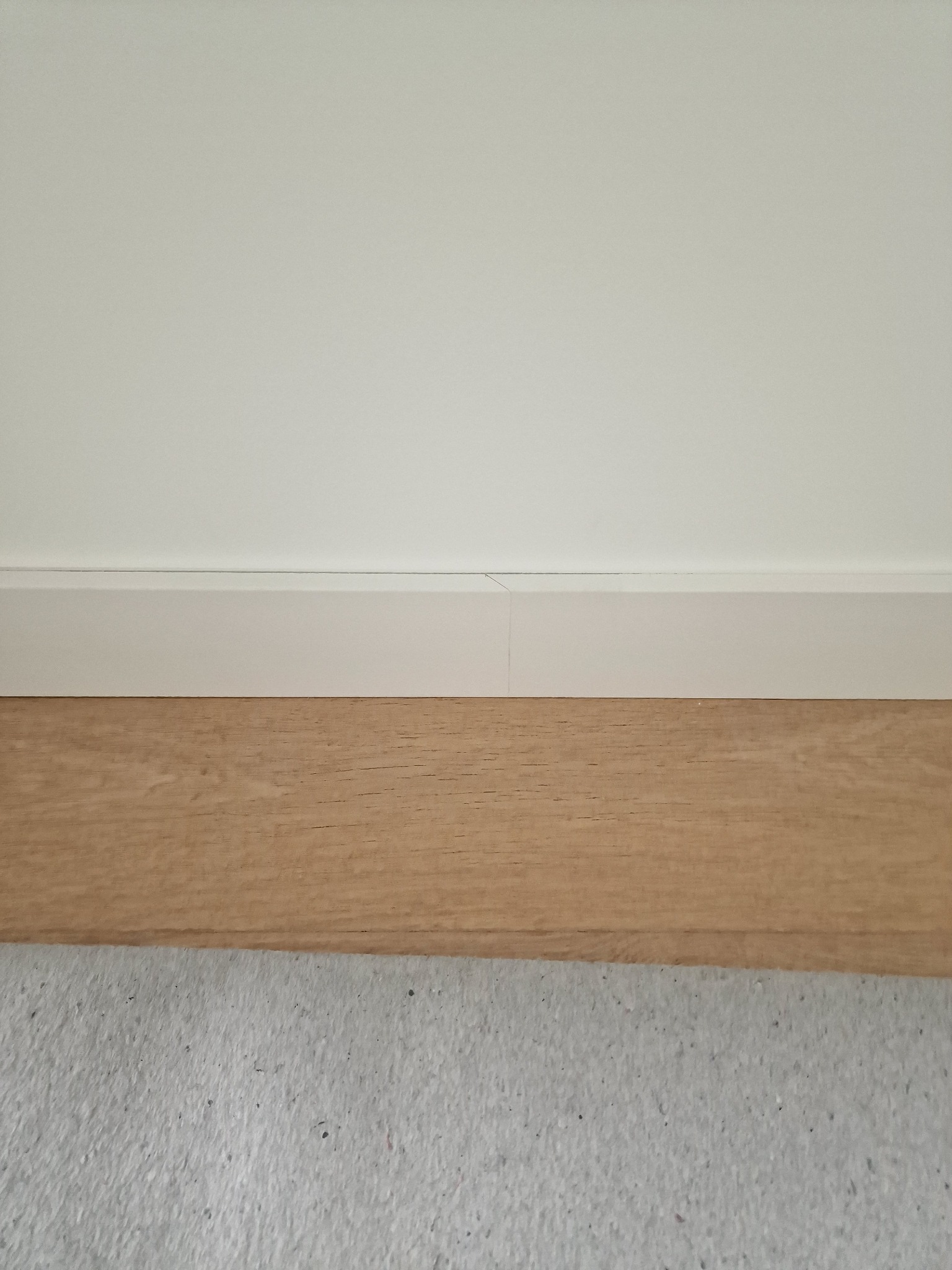 Jasny parkiet ułożony przy białej ścianie z białą listwą przypodłogową, widoczny fragment szarej wykładziny podłogowej.