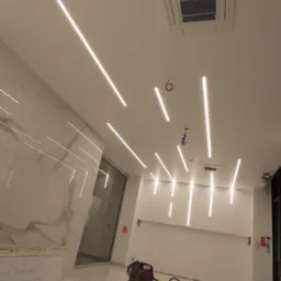 Nowoczesne oświetlenie liniowe LED na suficie w jasnym, przestronnym wnętrzu z marmurową ścianą i błyszczącą podłogą. Widoczny klimatyzator kasetonowy i odkurzacz.