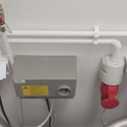 Tech-Net Kamil Machalski - Szare skrzynki elektryczne z czerwonymi gniazdami i białymi rurami na białej ścianie. Widoczne oznaczenia 105, 106, 108, 110 oraz naklejka ostrzegawcza.