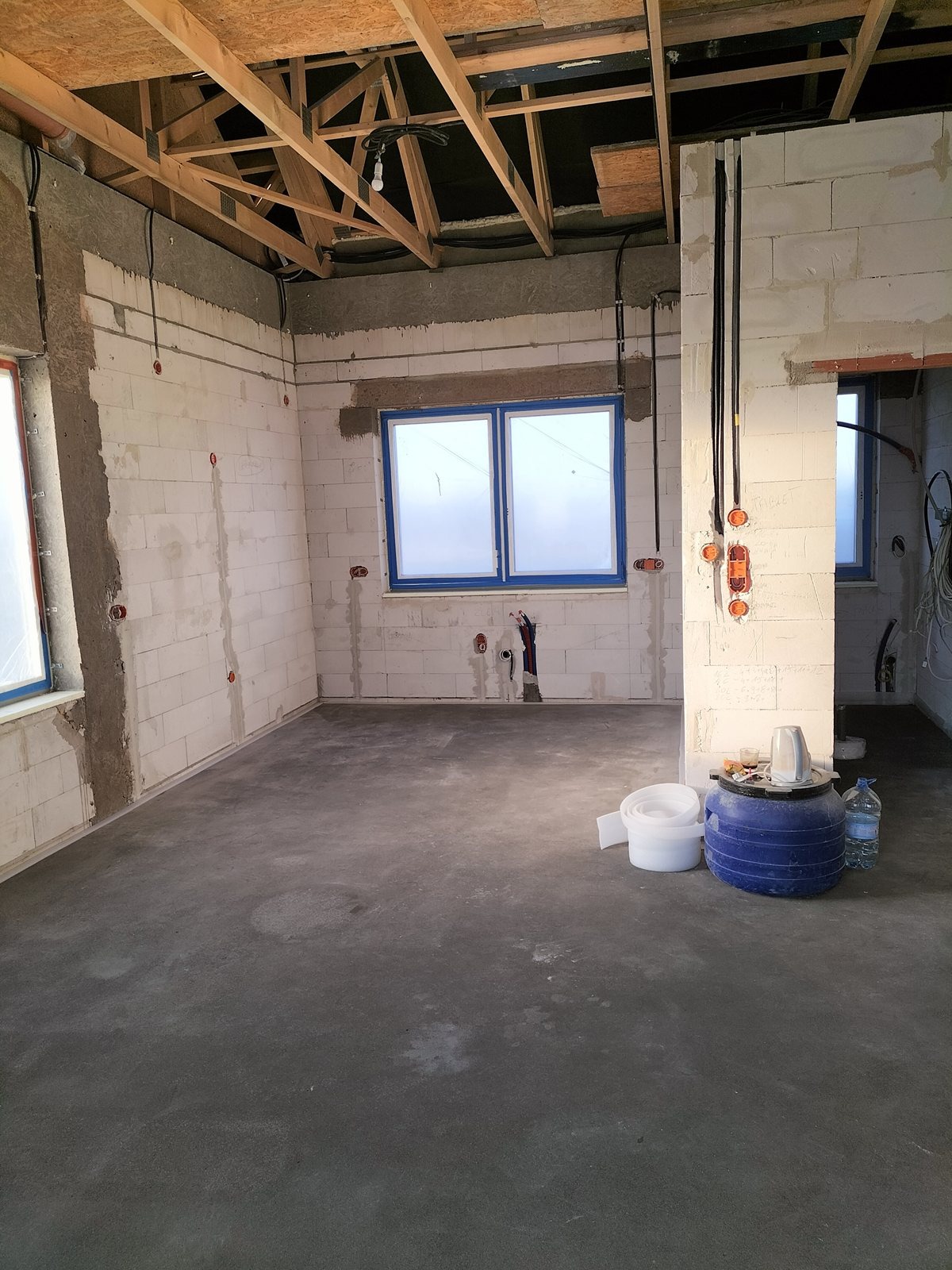 Surowy stan wnętrza: ściany z betonu komórkowego z widoczną instalacją elektryczną, w tle okna z niebieską ramą, na podłodze narzędzia i materiały budowlane.