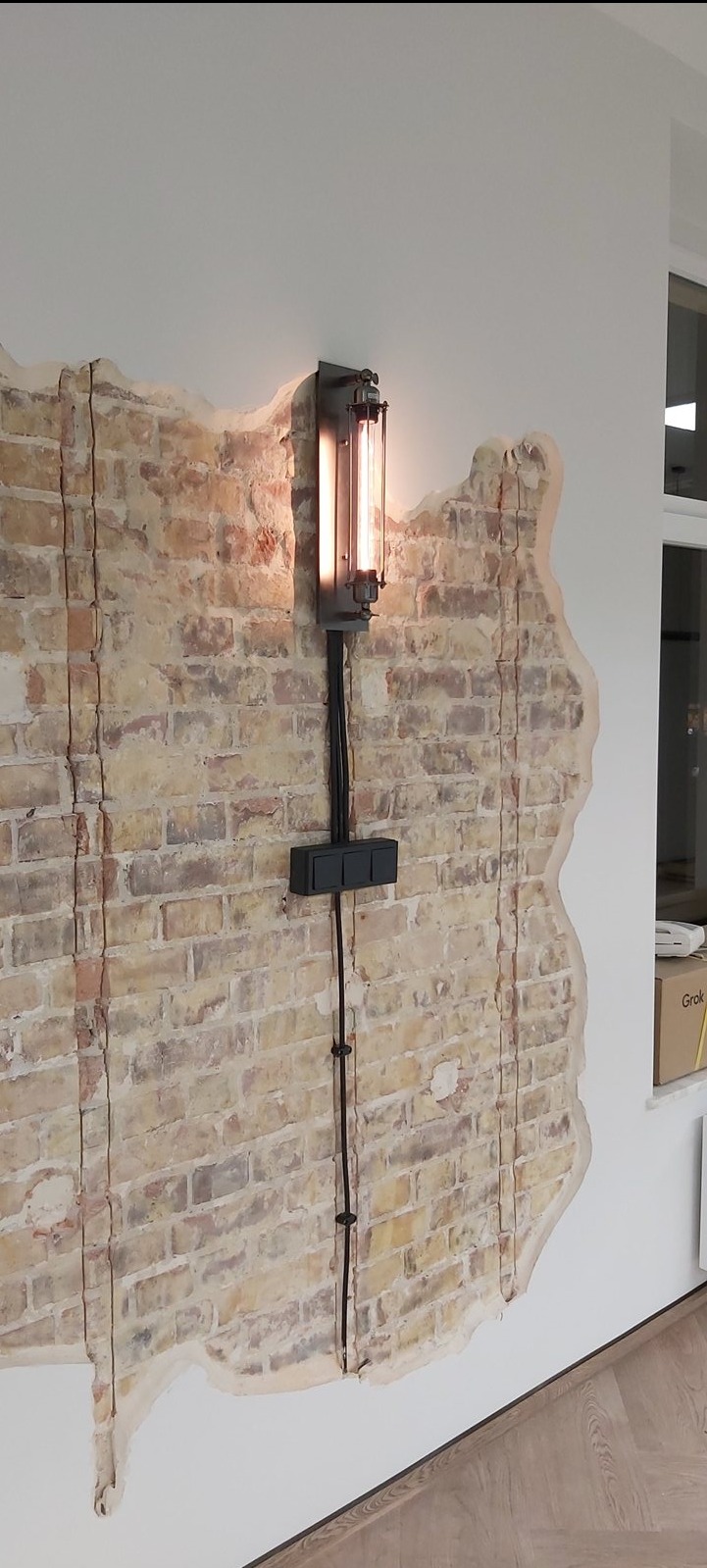 Industrialna lampa ścienna z widocznymi przewodami na tle odsłoniętej cegły i białej ściany, oświetlająca fragment wnętrza z drewnianą podłogą.