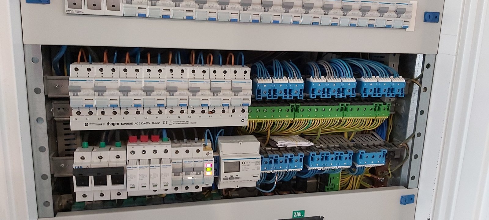 Szczegółowe ujęcie rozdzielnicy elektrycznej z bezpiecznikami Hager, okablowaniem w kolorze niebieskim, żółtym i zielonym oraz licznikiem energii. Widoczne oznaczenia L1, L2, L3, N.
