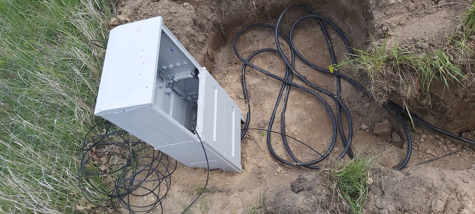 Otwarta, biała skrzynka elektryczna w wykopie ziemnym z wystającymi kablami. Widoczne elementy instalacji wewnątrz. Trawa i ziemia w tle.