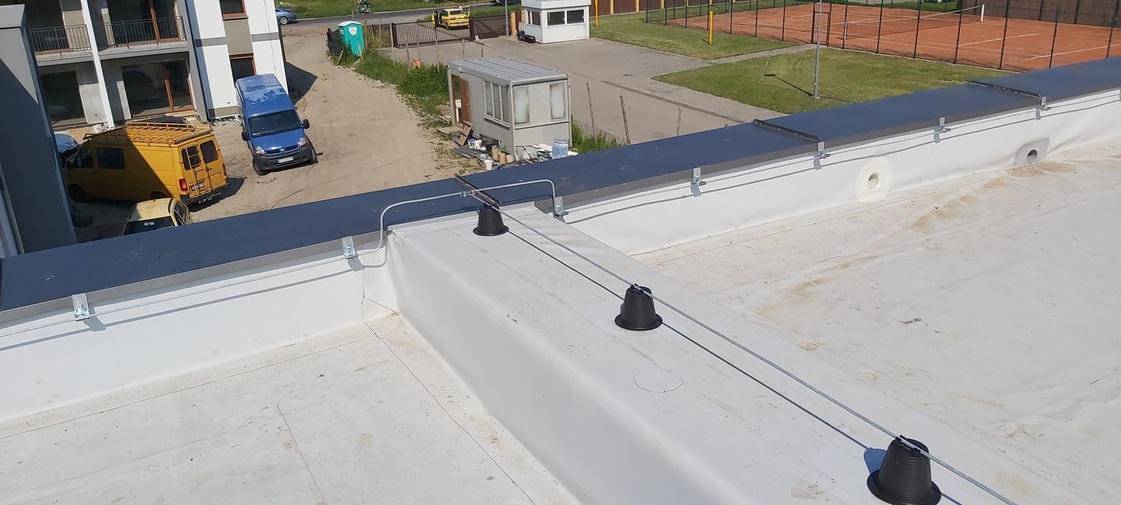 Instalacja odgromowa na białym dachu płaskim budynku. Widoczne czarne uchwyty i przewody. W tle budynki, parking z samochodami i kort tenisowy.