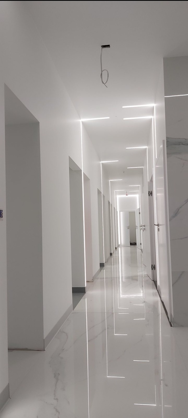 Długi, biały korytarz z nowoczesnym oświetleniem liniowym LED wzdłuż ścian i sufitu. Białe, błyszczące płytki podłogowe odbijają światło, tworząc efektowny wizualny efekt.