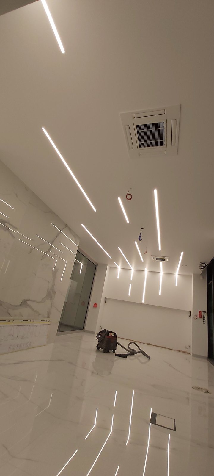 Nowoczesne oświetlenie liniowe LED na suficie w jasnym, przestronnym wnętrzu z marmurową ścianą i błyszczącą podłogą. Widoczny klimatyzator kasetonowy i odkurzacz.