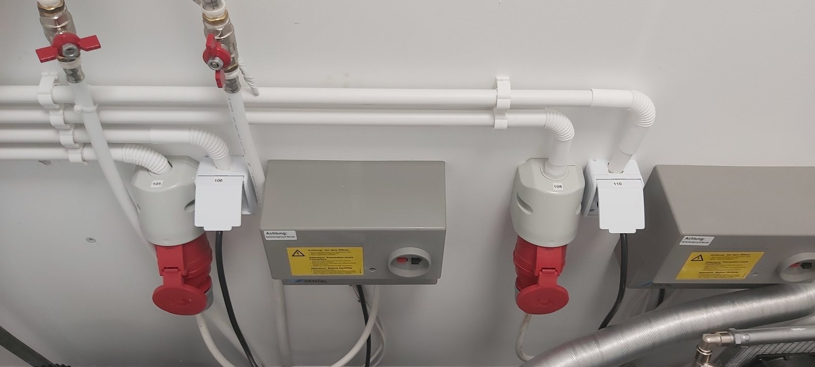 Szare skrzynki elektryczne z czerwonymi gniazdami i białymi rurami na białej ścianie. Widoczne oznaczenia 105, 106, 108, 110 oraz naklejka ostrzegawcza.