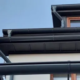 Eleganckie, grafitowe rynny zamontowane na dachu budynku z drewnianymi oknami, widoczne pod kątem z dołu na tle błękitnego nieba.