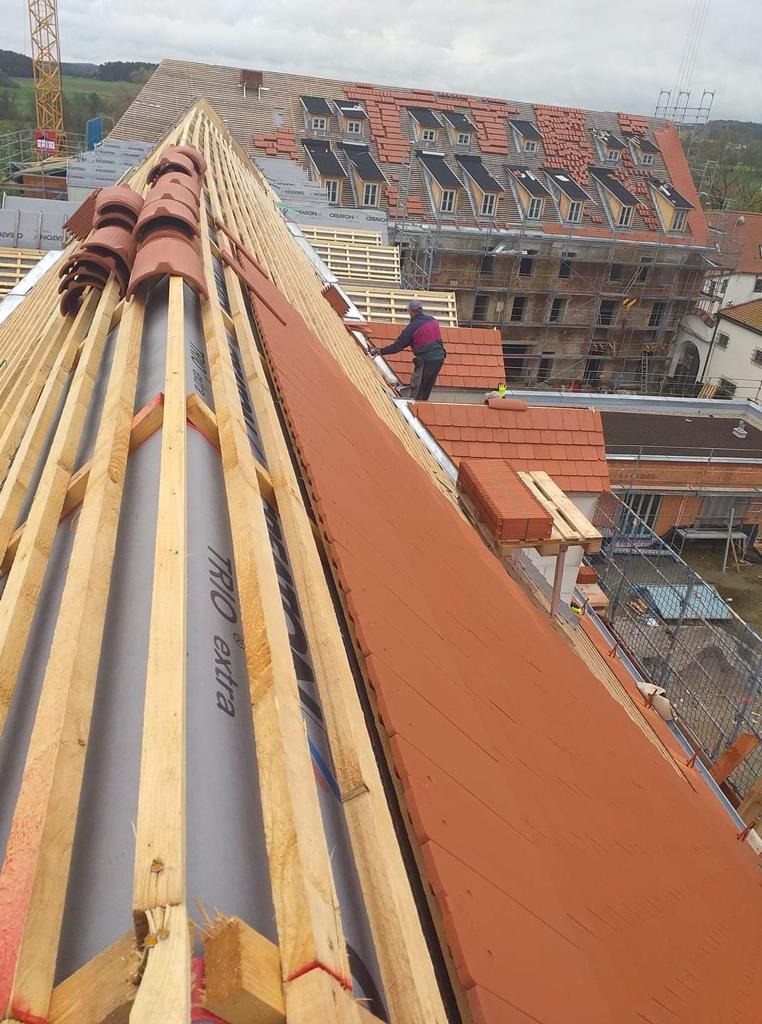 Prace dekarskie w toku: układanie czerwonej dachówki na drewnianej konstrukcji więźby dachowej, widok z góry na częściowo pokryty dach i pracującego dekarza, w tle budynek w rusztowaniach.