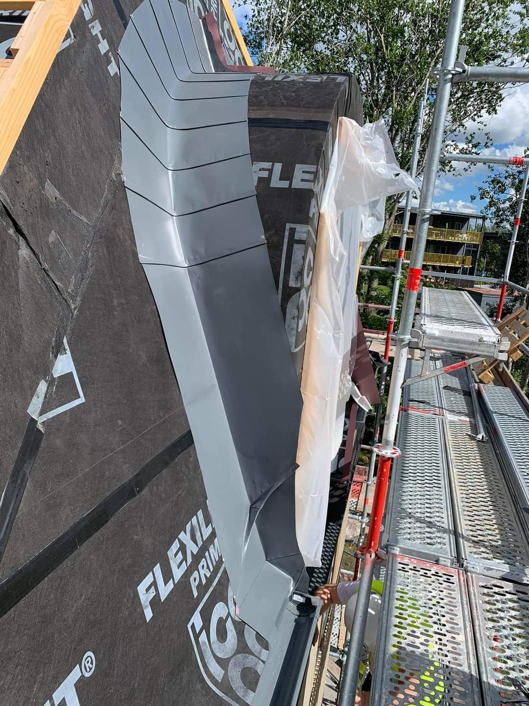 Szczegółowe ujęcie fragmentu dachu w trakcie montażu, widoczne czarne membrany dachowe, szare obróbki blacharskie na kominie oraz rusztowanie z pracownikiem w tle.