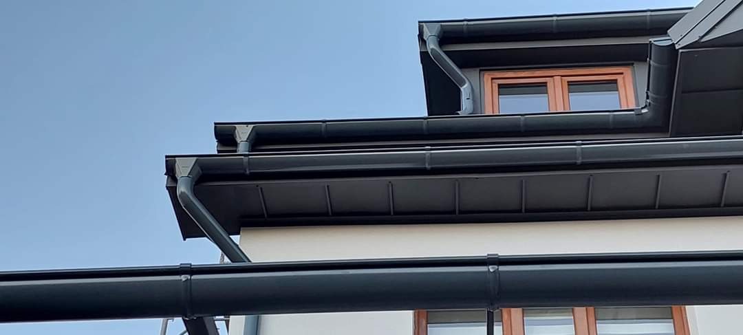 Eleganckie, grafitowe rynny zamontowane na dachu budynku z drewnianymi oknami, widoczne pod kątem z dołu na tle błękitnego nieba.