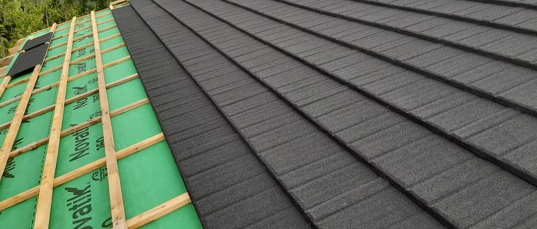 Fragment dachu w trakcie układania: widoczne drewniane łaty, zielona membrana i częściowo ułożona grafitowa dachówka.