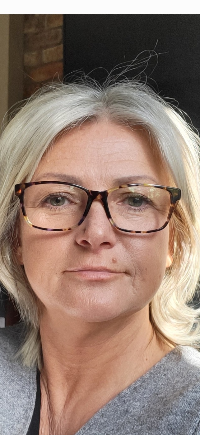 Portret kobiety z siwymi włosami i okularami w rogowej oprawce, patrzącej wprost w obiektyw, z widocznym znamieniem na policzku.