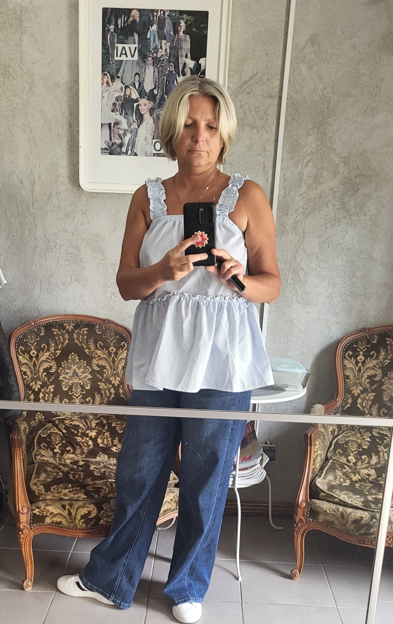 Osoba w bluzce w paski z falbankami i dżinsach szerokich nogawkach robi selfie w lustrze, w tle widoczne krzesła w stylu vintage i plakat z modą.