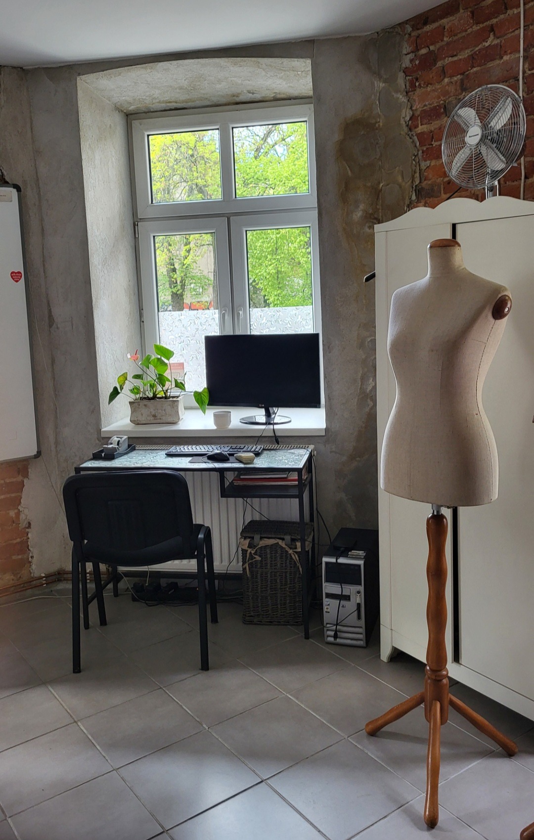 Pracownia krawiecka: Manekin krawiecki stoi obok biurka z komputerem, widok przez okno na zieleń drzew