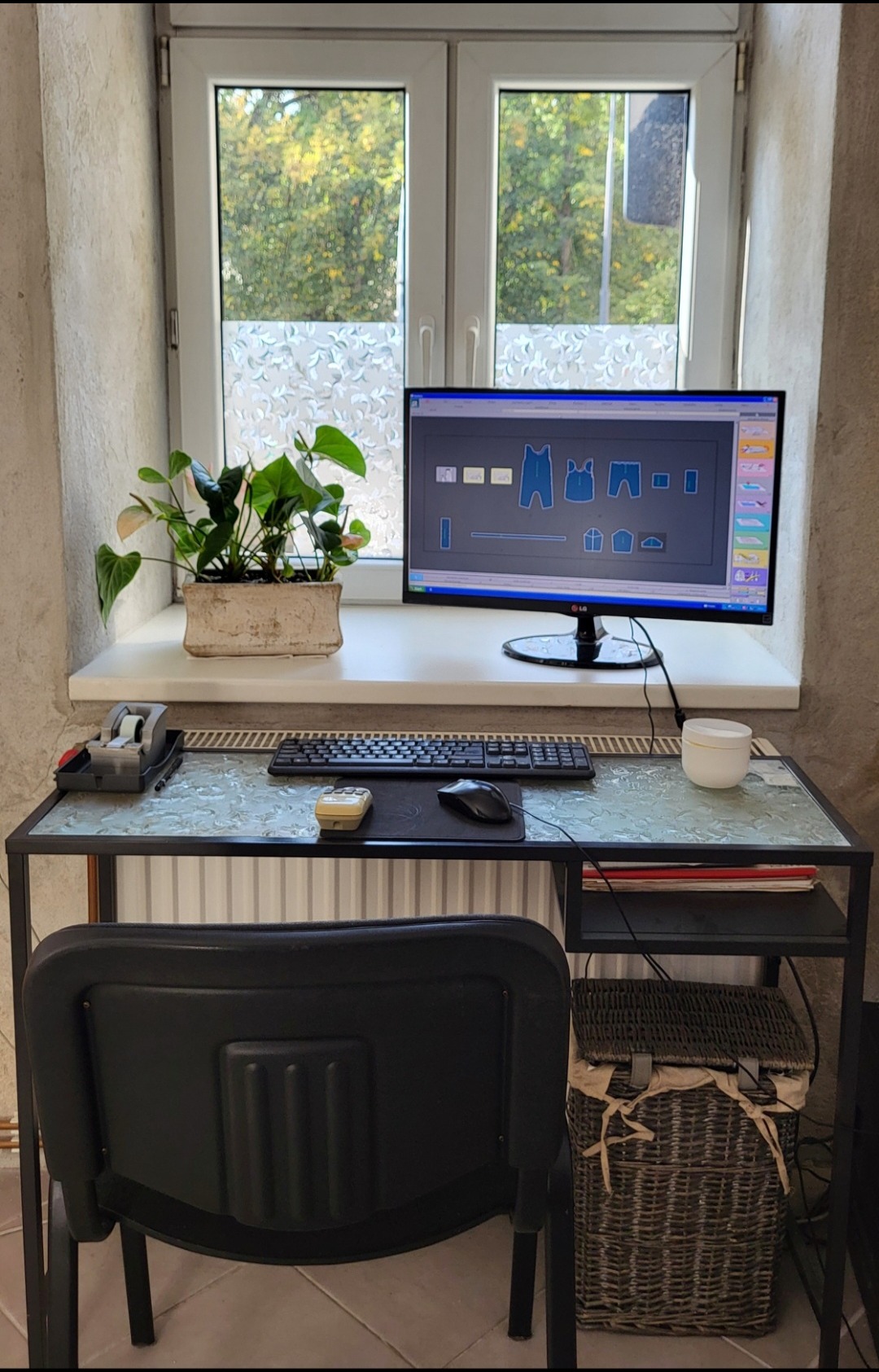 Stanowisko pracy wzorcowni odzieży z monitorem prezentującym projekt wykroju, rośliną doniczkową na parapecie i koszem wiklinowym pod biurkiem.