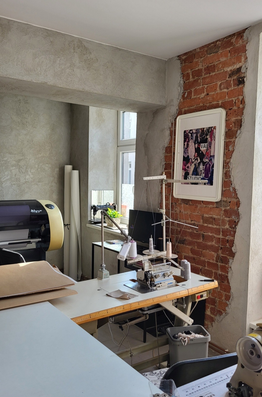 Wnętrze pracowni krawieckiej z maszyną do szycia JUKI na stole, drukarka wielkoformatowa w tle, ściana z cegły z plakatem w ramie.