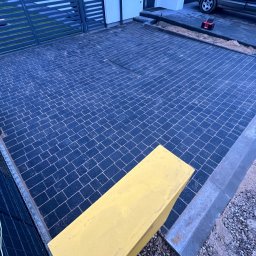 Adrian Pierzchot Paving Arts - Podjazd z ciemnej kostki brukowej ułożonej w wzór, z odpływem liniowym i żółtą donicą. Widoczne krawężniki najazdowe i brama w tle. Realizacja z wyczystką.