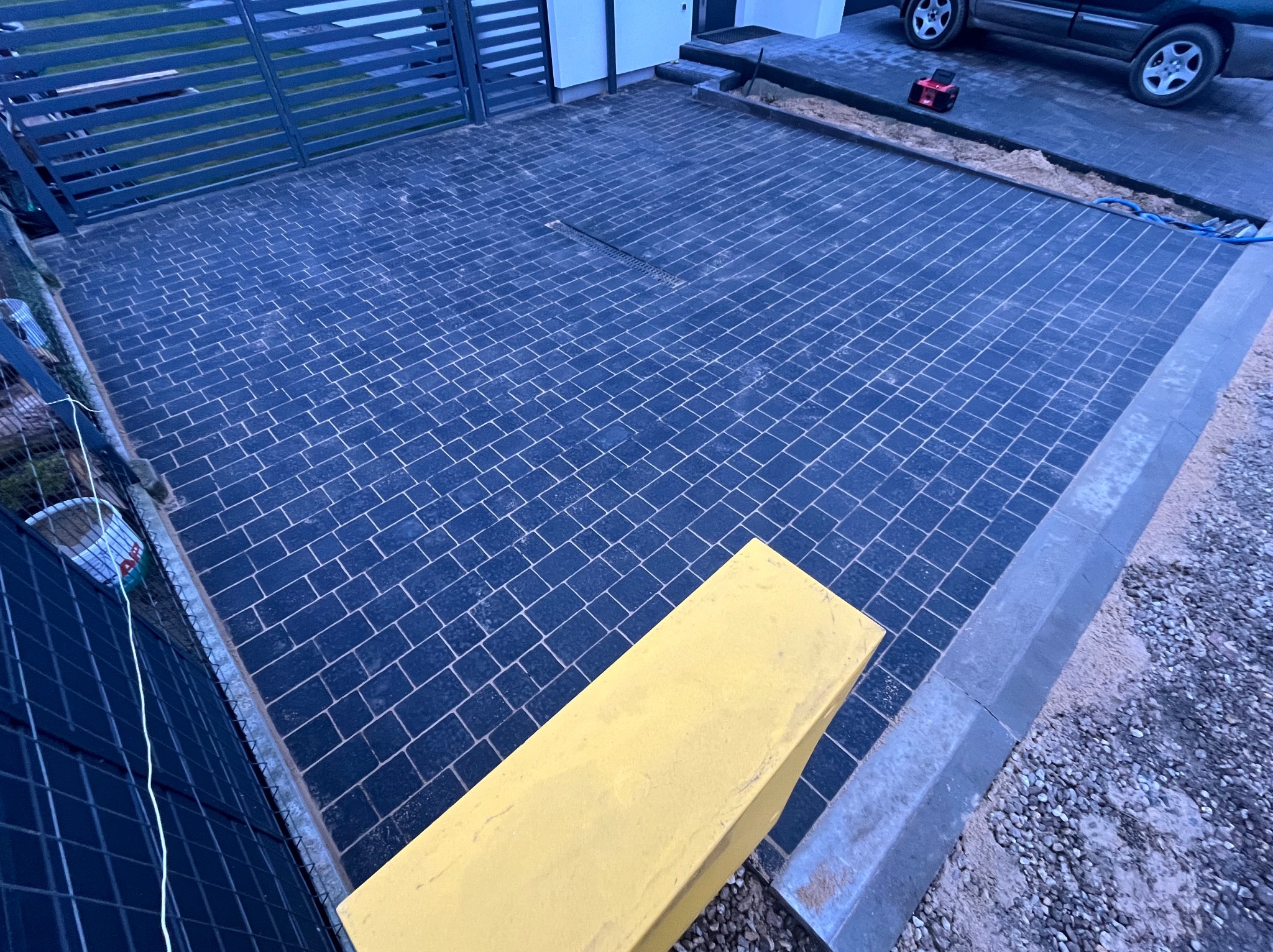 Podjazd z ciemnej kostki brukowej ułożonej w wzór, z odpływem liniowym i żółtą donicą. Widoczne krawężniki najazdowe i brama w tle. Realizacja z wyczystką.