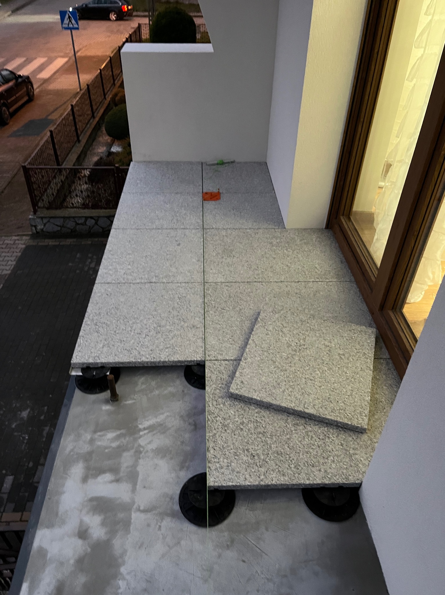 Montaż podłogi na balkonie z płyt imitujących granit, widoczne regulowane wsporniki. Częściowo ułożona podłoga z widokiem na ulicę i budynek.