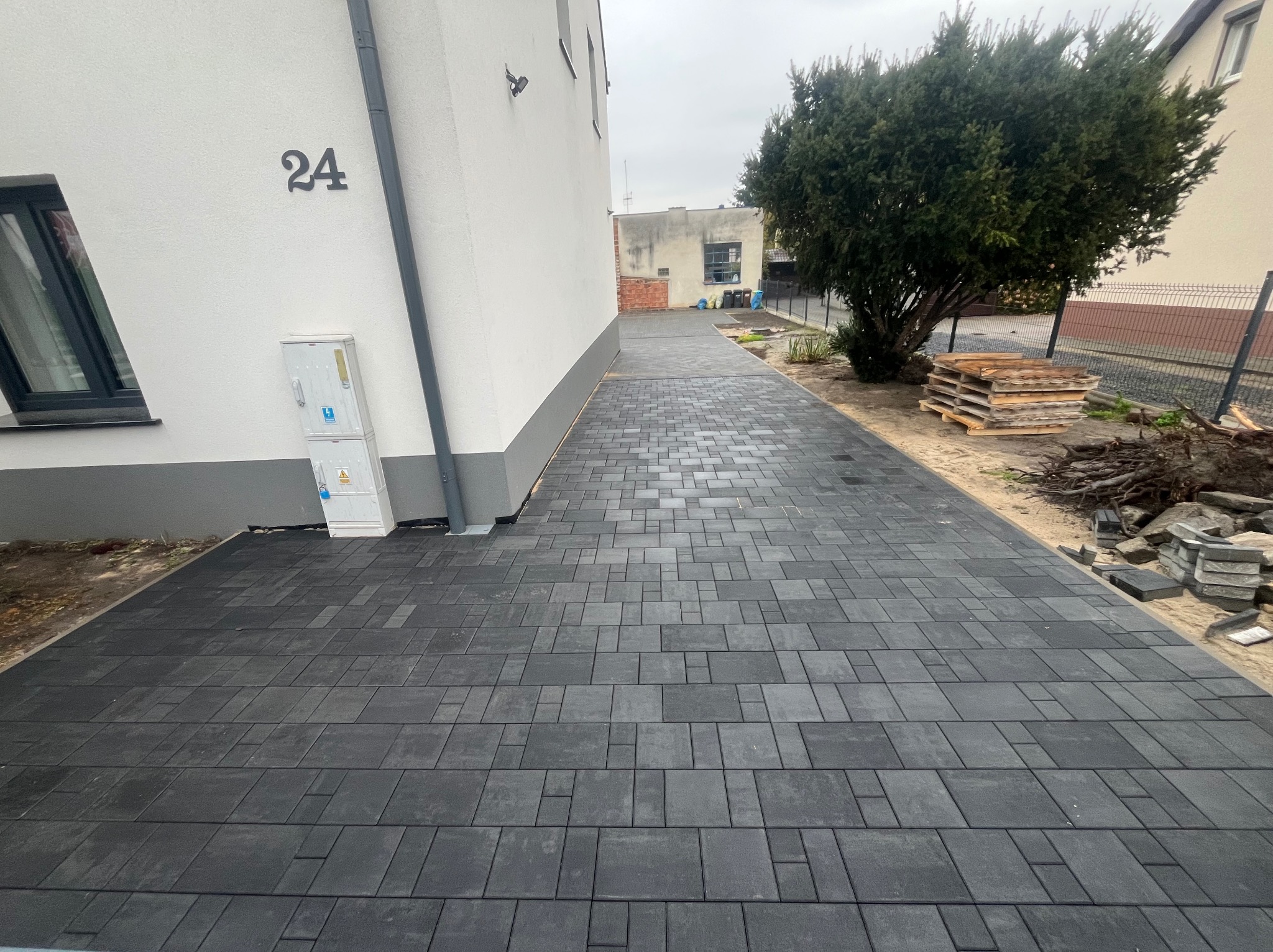 Ułożona, grafitowa kostka brukowa prowadząca do budynku z numerem 24. Widoczne palety i resztki materiałów budowlanych. Ujęcie z perspektywy.