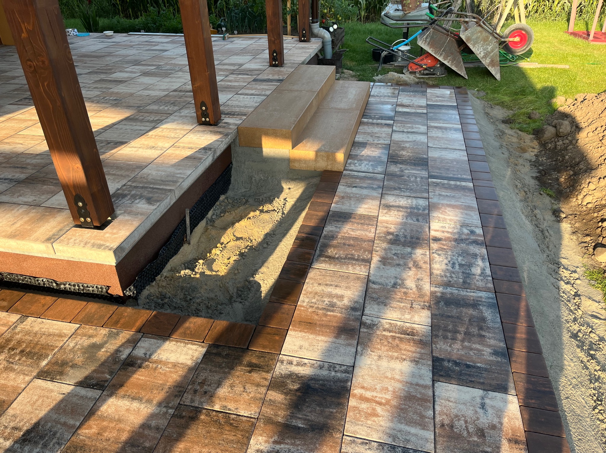 Układanie kostki: taras z brązowo-beżowej kostki, widoczne etapy prac przy schodach i obrzeżach. W tle narzędzia i trawnik.