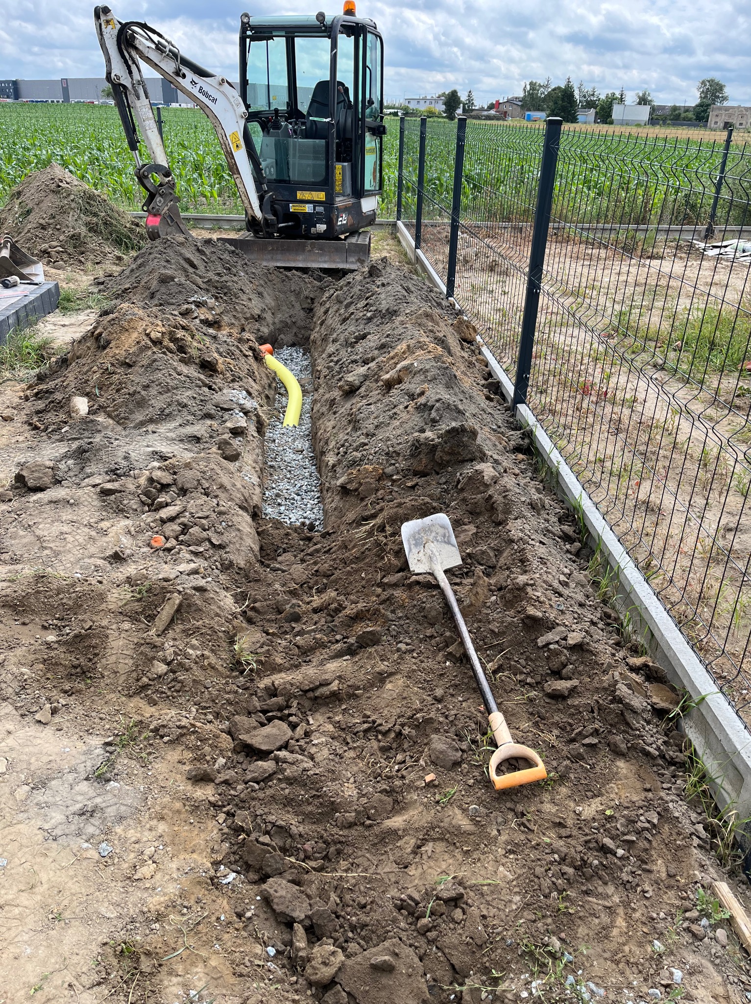 Wykopany rów z żółtą rurą drenażową i warstwą żwiru, obok leży łopata. W tle minikoparka i ogrodzenie panelowe. Prace ziemne na działce z uprawami.