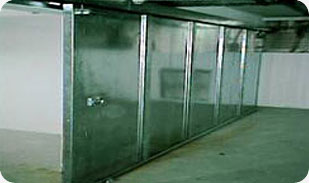 Zamknięte, metalowe drzwi segmentowe w hali przemysłowej, widoczne łączenia segmentów, szara kolorystyka, oświetlenie górne.