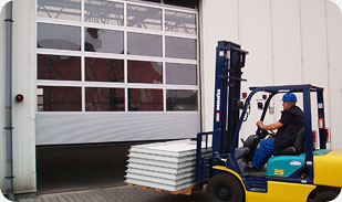 Wózek widłowy z operatorem transportuje panele segmentowe przed częściowo otwartą bramą garażową przemysłową z przezroczystymi sekcjami, widoczny fragment elewacji budynku.