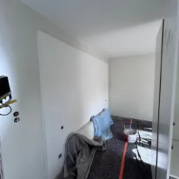 Pokój w trakcie odświeżania, ściany pomalowane na biało, zabezpieczona podłoga, widoczne puszki z farbą i narzędzia malarskie, jeszcze nie posprzątane po pracy.