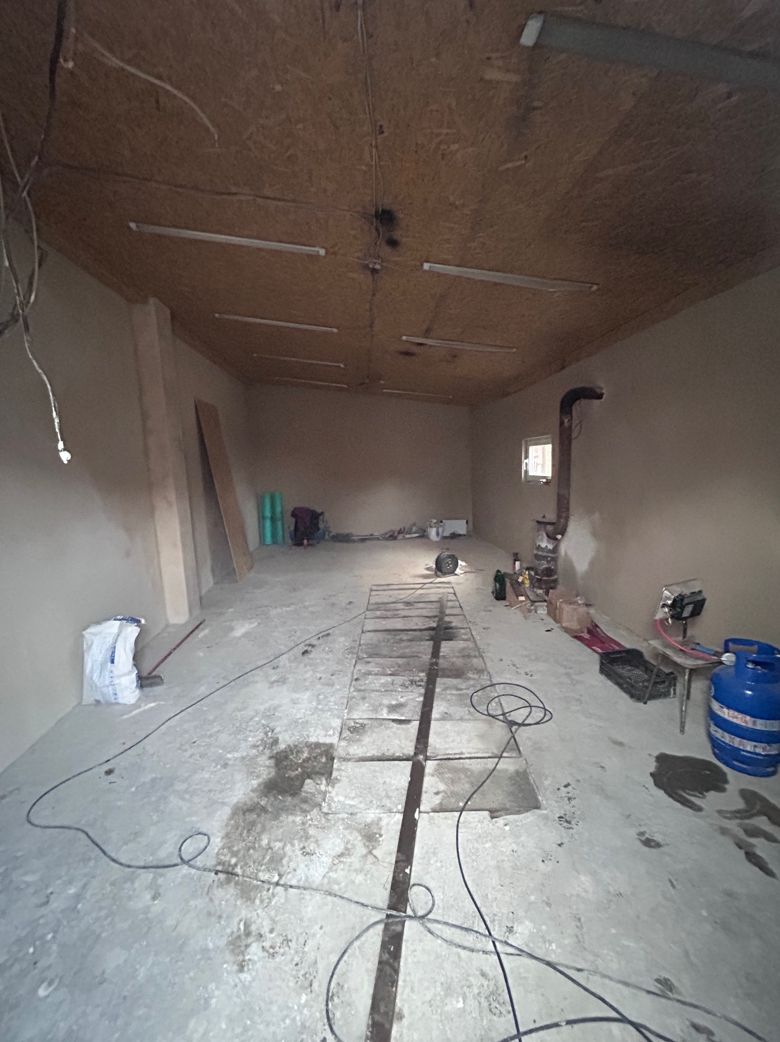 Wnętrze remontowanego garażu z widocznymi świeżo otynkowanymi ścianami, drewnianym sufitem z lampami, piecem typu koza i butlą gazową w rogu.
