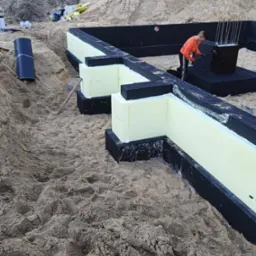 Fundamenty domu w trakcie budowy: bloczki styropianowe, czarna hydroizolacja, zbrojenie i pracownik zabezpieczający konstrukcję.