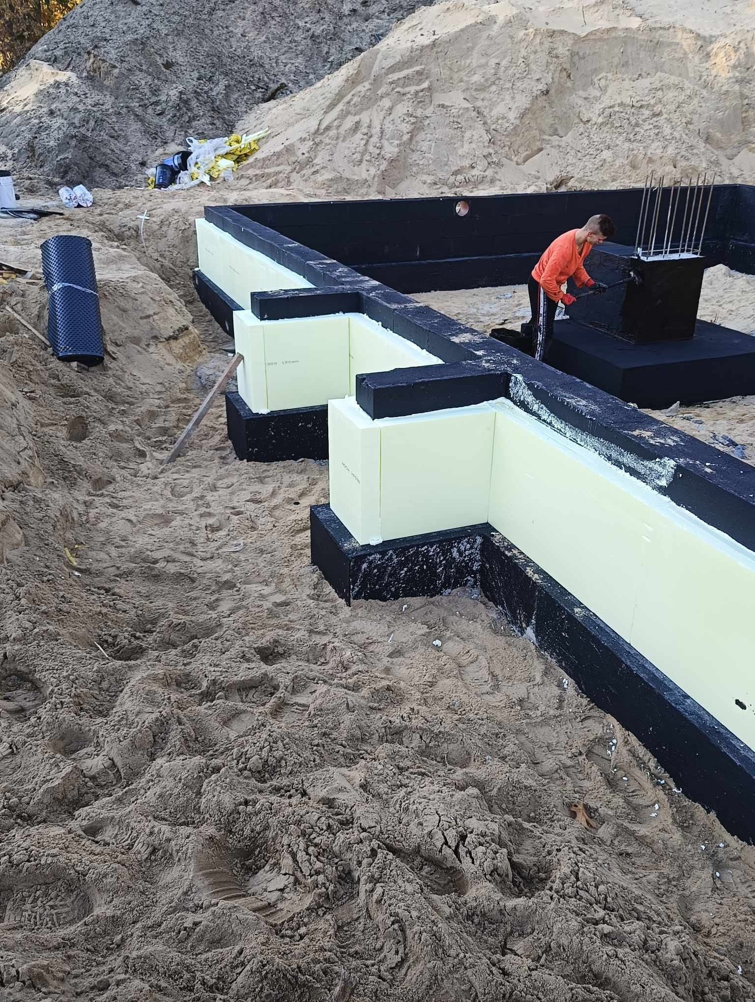 Fundamenty domu w trakcie budowy: bloczki styropianowe, czarna hydroizolacja, zbrojenie i pracownik zabezpieczający konstrukcję.