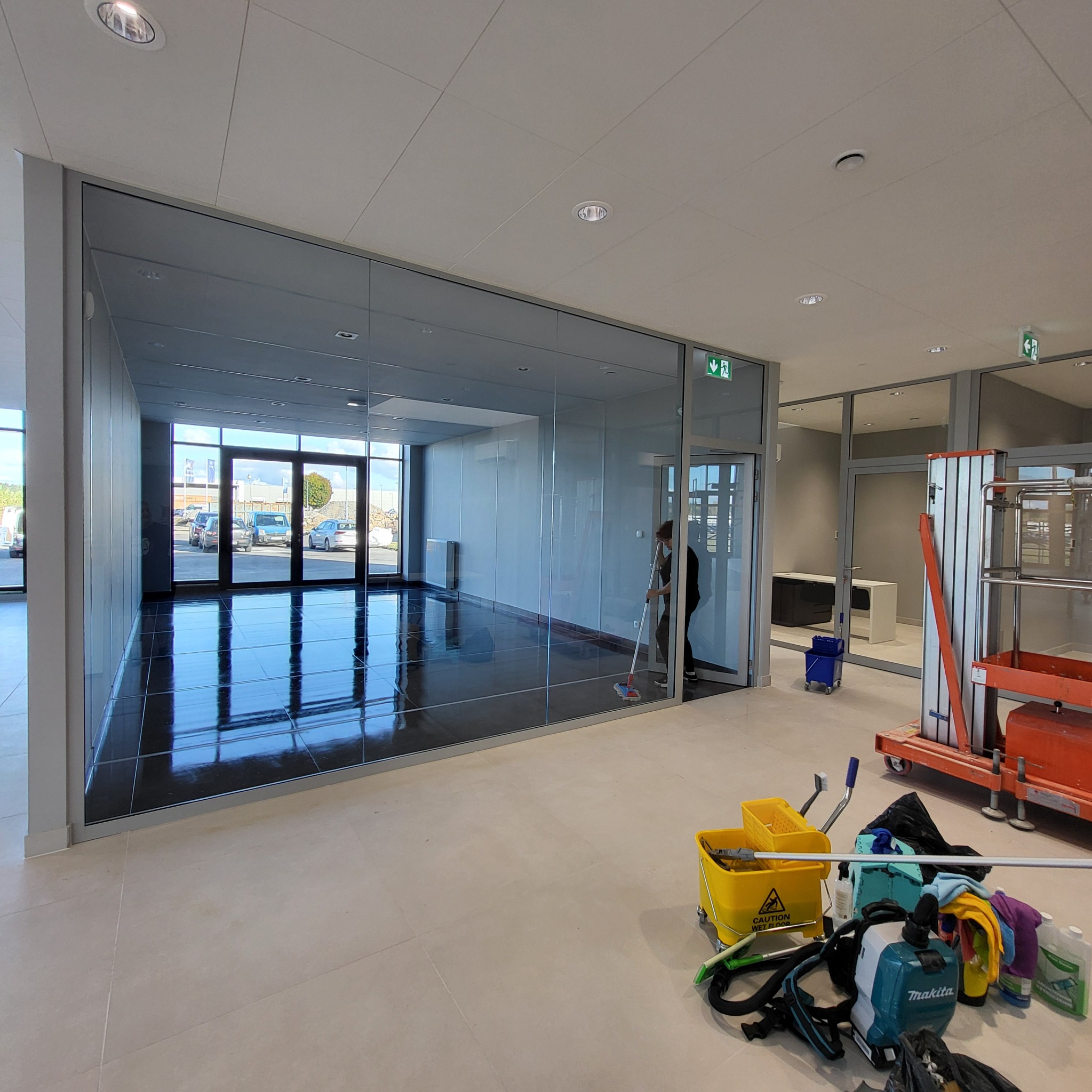 Pracownik w czarnej odzieży myje podłogę mopem w nowoczesnym biurze z przeszklonymi ścianami; widoczne wiadro z ostrzeżeniem o mokrej podłodze, odkurzacz i podnośnik.