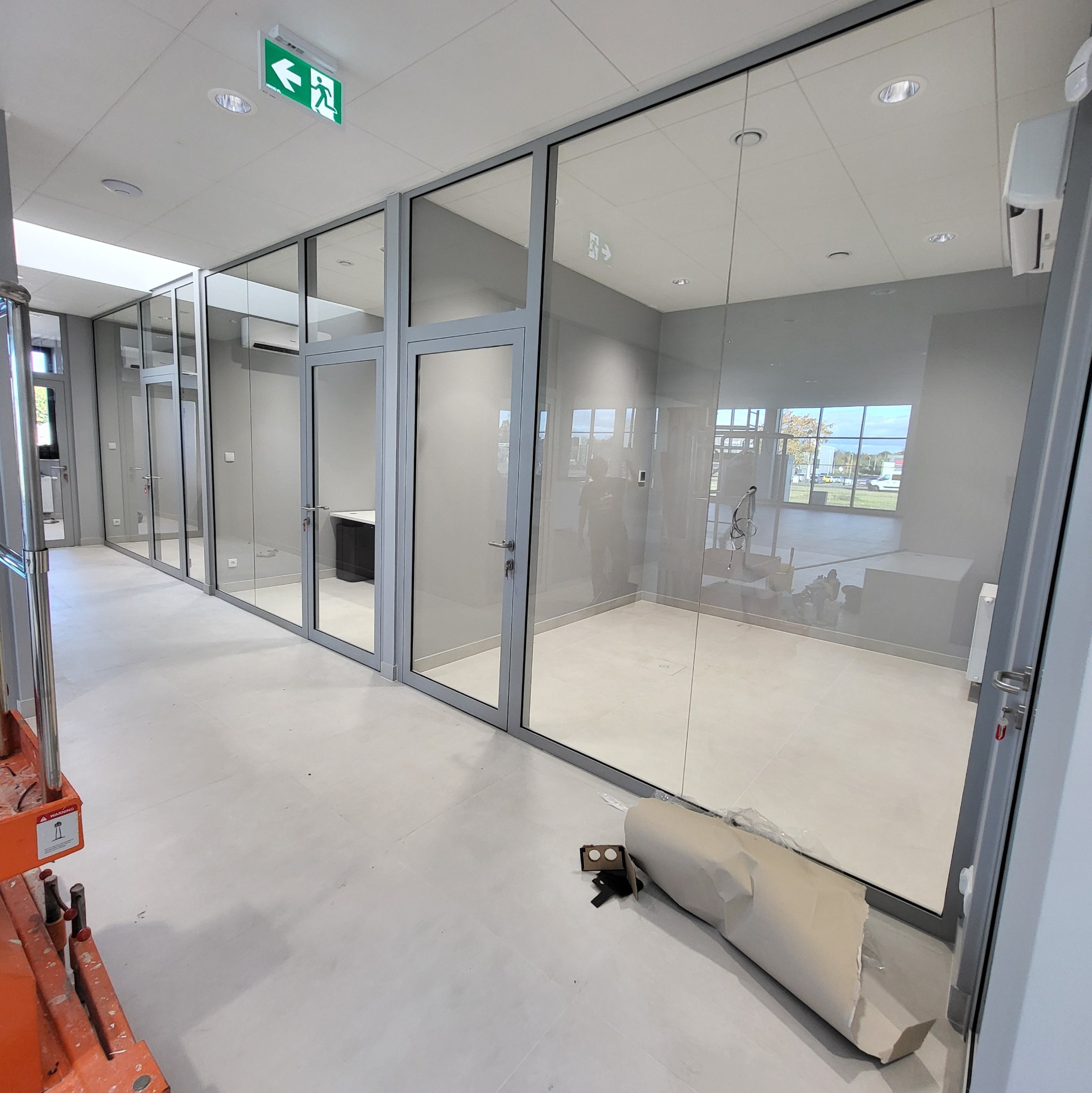 Wnętrze biura z przeszklonymi ścianami i drzwiami, widoczny podnośnik i resztki po pracach wykończeniowych na podłodze.