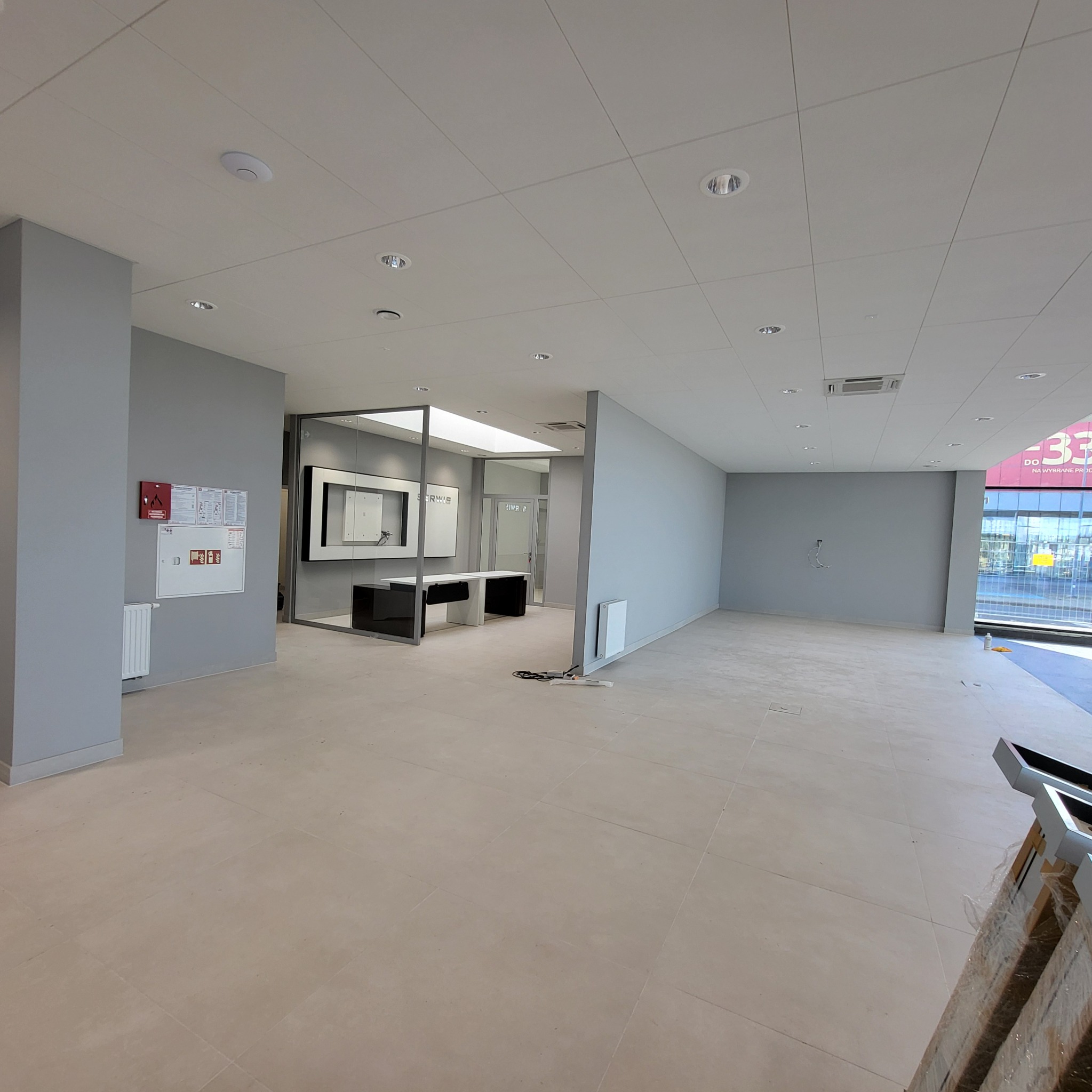 Pusta, jasna przestrzeń biurowa z szarymi ścianami, beżową podłogą z płytek, sufitem kasetonowym i minimalistycznym wyposażeniem, widoczne elementy instalacji elektrycznej na ścianie.