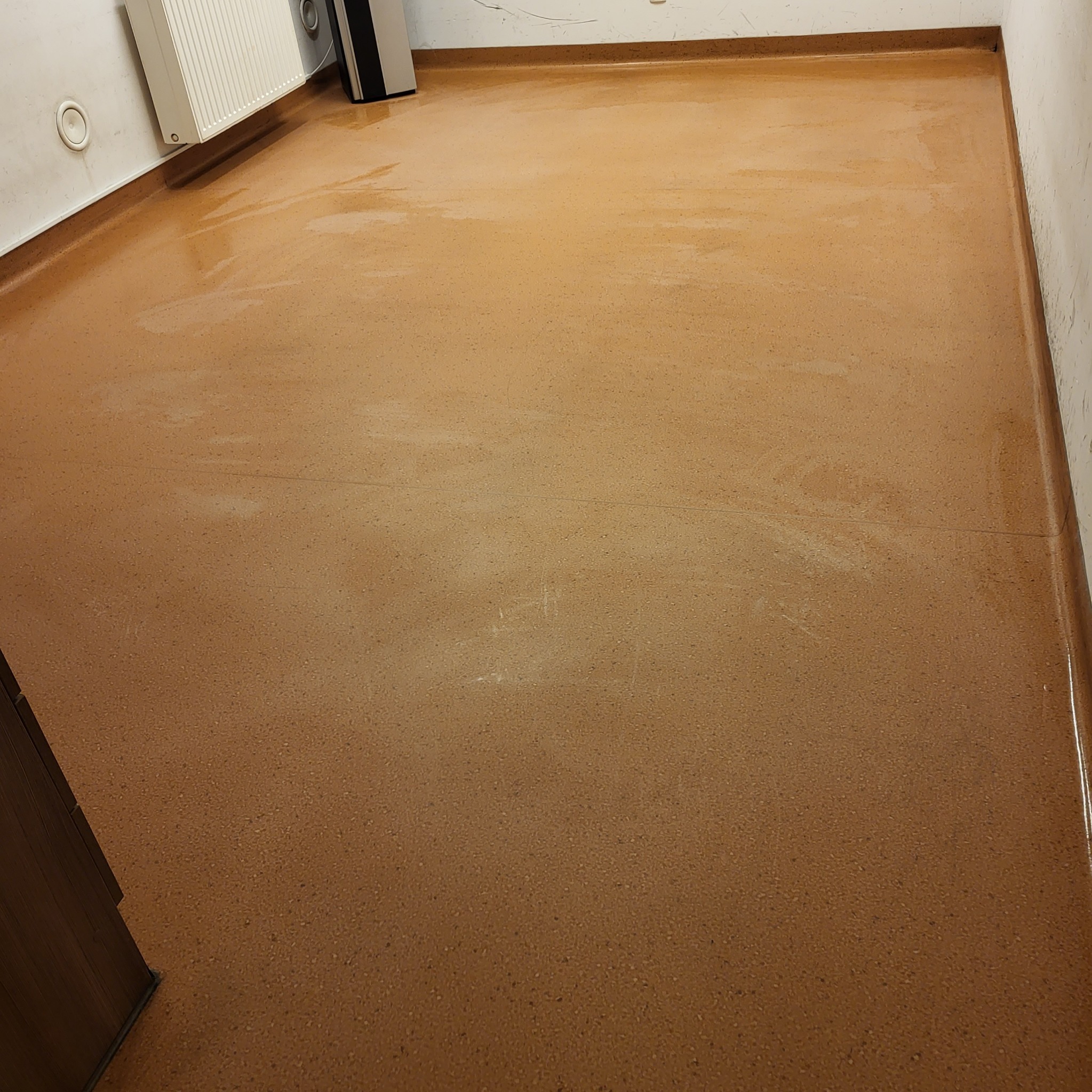 Błyszcząca, brązowa podłoga z drobnymi plamami, prawdopodobnie po umyciu, widoczny fragment ściany z grzejnikiem i listwą przypodłogową.