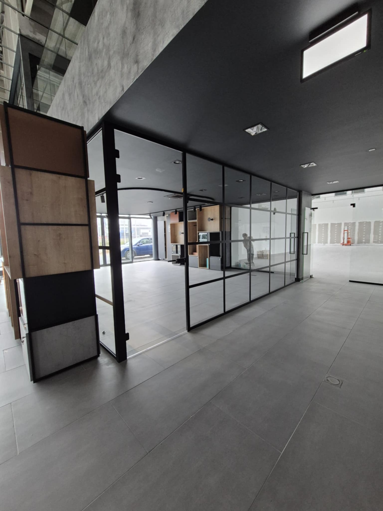 Wnętrze biura z przeszklonymi ścianami działowymi, widoczna osoba w tle i szara podłoga z płytek.